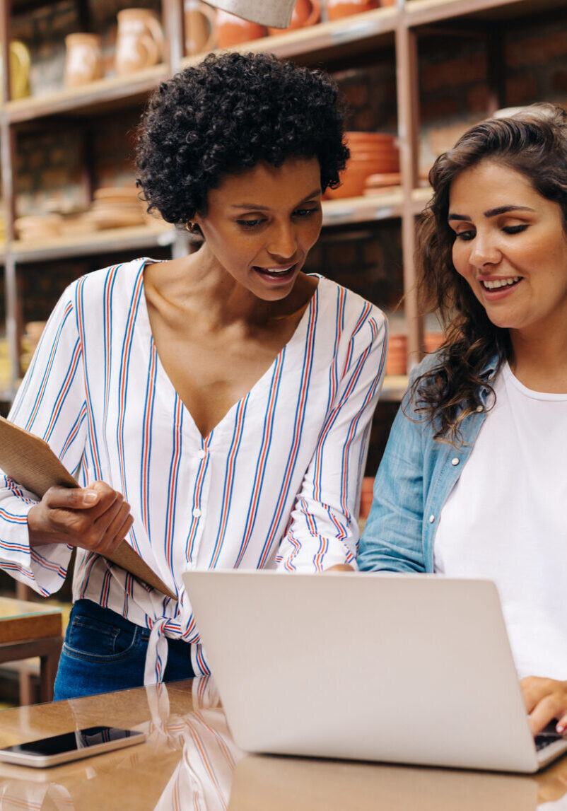 Two young shop owners using a laptop while working in a ceramic store. Female entrepreneurs managing online orders on their website. Young businesswomen running a creative small business together.